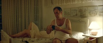 Movie still from “Dogtooth” (2009), directed by Yorgos Lanthimos – A man sitting on a bed playing a video game; Medium shot, Low angle