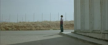 Movie still from “Dogtooth” (2009), directed by Yorgos Lanthimos – A person standing on a sidewalk near a fence; Extreme Wide shot, Low angle