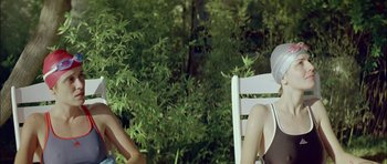 Movie still from “Dogtooth” (2009), directed by Yorgos Lanthimos – Two people sitting on a white chair in front of some trees; Medium shot, Low angle