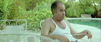 Movie still from “Dogtooth” (2009), directed by Yorgos Lanthimos – A man sitting on a chair near a swimming pool; Medium shot, Low angle