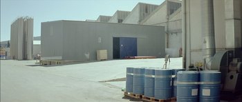 Movie still from “Dogtooth” (2009), directed by Yorgos Lanthimos – Several barrels of blue liquid sitting on top of pallets in front of an industrial building; Extreme Wide shot, Low angle
