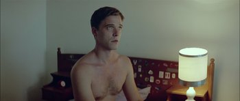 Movie still from “Dogtooth” (2009), directed by Yorgos Lanthimos – A man sitting on top of a bed in a room; Medium shot, High angle