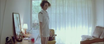 Movie still from “Dogtooth” (2009), directed by Yorgos Lanthimos – A woman standing in front of a yellow chair in front of a window; Medium shot, Low angle