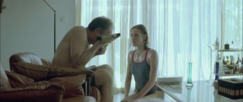 Movie still from “Dogtooth” (2009), directed by Yorgos Lanthimos – A man taking a picture of a woman sitting on the ground; Medium shot, Over the shoulder angle