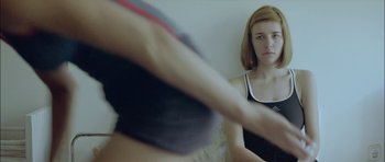 Movie still from “Dogtooth” (2009), directed by Yorgos Lanthimos – A woman looking into a mirror while a man holds his arm up; Medium shot, Over the shoulder angle