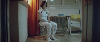 Movie still from “Dogtooth” (2009), directed by Yorgos Lanthimos – A woman sitting on a chair in front of a table; Medium shot, Over the shoulder angle