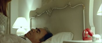 Movie still from “Dogtooth” (2009), directed by Yorgos Lanthimos – A man laying in a bed next to an ornate headboard; Close Up shot, Low angle