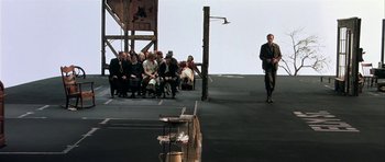 Movie still from “Dogville” (2003), directed by Lars von Trier – A group of people sitting and standing on a stage; Wide shot, High angle