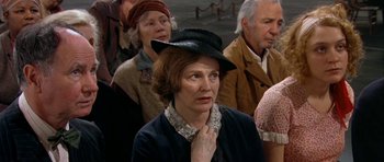 Movie still from “Dogville” (2003), directed by Lars von Trier – An older woman wearing a black hat in front of a group of older people; Medium shot, High angle