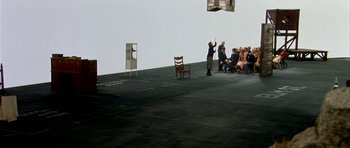 Movie still from “Dogville” (2003), directed by Lars von Trier – A group of people sitting on the ground in front of a white wall; Extreme Wide shot, High angle