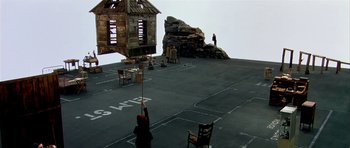 Movie still from “Dogville” (2003), directed by Lars von Trier – A person standing on a deck with a chair and a chair; Extreme Wide shot, High angle