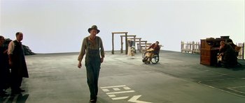 Movie still from “Dogville” (2003), directed by Lars von Trier – A man walking in front of another man in a wheel chair; Wide shot, High angle