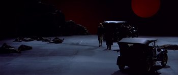 Movie still from “Dogville” (2003), directed by Lars von Trier – Two people standing in a parking lot next to a car; Extreme Wide shot, High angle