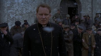 Movie still from “Dominion: Prequel to the Exorcist” (2005), directed by Paul Schrader – A man wearing a priest's outfit standing in front of a group of people; Close Up shot, Over the shoulder angle