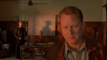 Movie still from “Dominion: Prequel to the Exorcist” (2005), directed by Paul Schrader – A man standing in front of a table in a room; Close Up shot, Over the shoulder angle