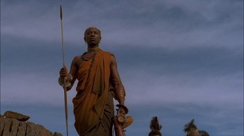 Movie still from “Dominion: Prequel to the Exorcist” (2005), directed by Paul Schrader – A man in an orange robe holding a spear; Medium shot, Low angle