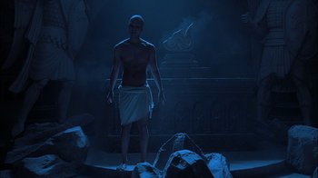 Movie still from “Dominion: Prequel to the Exorcist” (2005), directed by Paul Schrader – A man standing in a dark room with rocks and a statue; Wide shot, Low angle