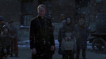 Movie still from “Dominion: Prequel to the Exorcist” (2005), directed by Paul Schrader – A man standing in front of a group of people in the snow; Medium shot, Over the shoulder angle