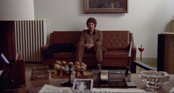 Movie still from “Don't Look Now” (1973), directed by Nicolas Roeg – A man sitting on top of a brown couch in a living room; Wide shot, Low angle