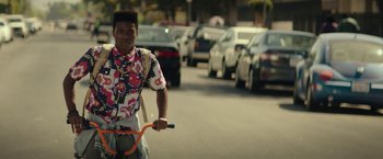 Movie still from “Dope” (2015), directed by Rick Famuyiwa – A man riding a bike down a street next to parked cars; Medium shot, Over the shoulder angle