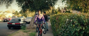 Movie still from “Dope” (2015), directed by Rick Famuyiwa – A man riding a bike down a sidewalk next to a tree; Wide shot, Over the shoulder angle