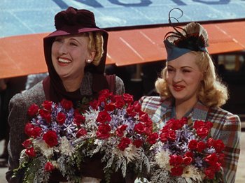 Movie still from “Down Argentine Way” (1940), directed by Irving Cummings – Two women in hats are smiling while holding flowers; Medium shot, Low angle