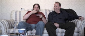 Movie still from “Down Terrace” (2009), directed by Ben Wheatley – Two people sitting on a couch eating and watching television; Medium shot, Over the shoulder angle