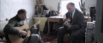 Movie still from “Down Terrace” (2009), directed by Ben Wheatley – A man sitting in front of another man holding a guitar; Medium shot, Low angle