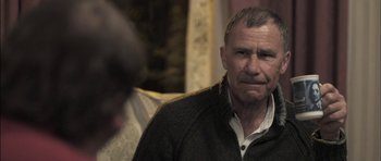 Movie still from “Down Terrace” (2009), directed by Ben Wheatley – An older man sitting on a couch looking at the camera; Close Up shot, Over the shoulder angle