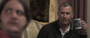 Movie still from “Down Terrace” (2009), directed by Ben Wheatley – An older man sitting on a couch in a living room; Close Up shot, Over the shoulder angle