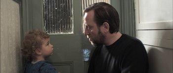 Movie still from “Down Terrace” (2009), directed by Ben Wheatley – A man with long hair and a goatee is looking at a child; Close Up shot, Over the shoulder angle