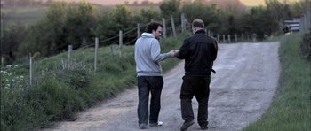 Movie still from “Down Terrace” (2009), directed by Ben Wheatley – Two men standing next to each other on a dirt road; Medium shot, Over the shoulder angle