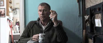 Movie still from “Down Terrace” (2009), directed by Ben Wheatley – A man sitting in front of a coffee mug holding a cookie; Close Up shot, Over the shoulder angle