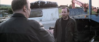 Movie still from “Down Terrace” (2009), directed by Ben Wheatley – A man holding a gun in front of another man in front of an old truck; Medium shot, Over the shoulder angle