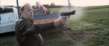 Movie still from “Down Terrace” (2009), directed by Ben Wheatley – A man holding a gun in a field; Medium shot, Low angle