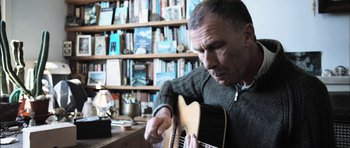 Movie still from “Down Terrace” (2009), directed by Ben Wheatley – A man playing a guitar in front of a book shelf; Close Up shot, Low angle