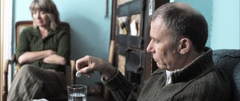 Movie still from “Down Terrace” (2009), directed by Ben Wheatley – An older man sitting at a table with a glass of water; Close Up shot, Over the shoulder angle