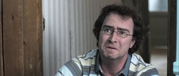 Movie still from “Down Terrace” (2009), directed by Ben Wheatley – A person wearing glasses and a striped shirt; Close Up shot, Over the shoulder angle