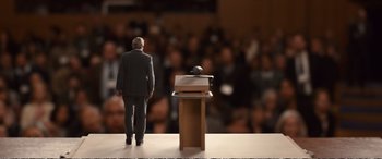 Movie still from “Downsizing” (2017), directed by Alexander Payne – A man standing at a podium with a microphone; Wide shot, High angle
