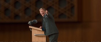 Movie still from “Downsizing” (2017), directed by Alexander Payne – A man giving a speech at a podium with a microphone; Medium shot, Over the shoulder angle