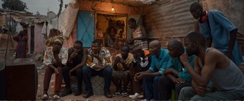Movie still from “Downsizing” (2017), directed by Alexander Payne – A group of men sitting on a chair in front of a brick building; Medium shot, Over the shoulder angle