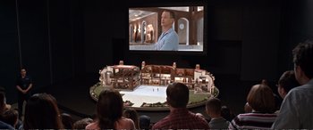 Movie still from “Downsizing” (2017), directed by Alexander Payne – A group of people sitting in front of a screen; Wide shot, High angle