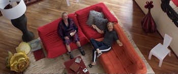 Movie still from “Downsizing” (2017), directed by Alexander Payne – Two men sitting on a red couch in a living room; Wide shot, Overhead angle
