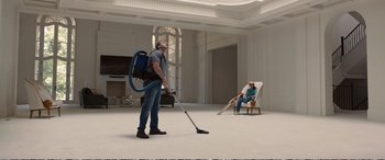 Movie still from “Downsizing” (2017), directed by Alexander Payne – A man is vacuuming while a man sits in a chair; Wide shot, Low angle