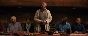 Movie still from “Downsizing” (2017), directed by Alexander Payne – An older man standing in front of a group of people at a dinner table; Medium shot, Over the shoulder angle