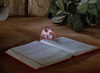 Movie still from “Dr. Cyclops” (1940), directed by Ernest B. Schoedsack – A man sitting on top of an open book next to a cactus plant; Wide shot, High angle