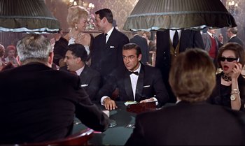 Movie still from “Dr. No” (1962), directed by Terence Young – A man in a tuxedo sitting at a table with other people; Medium shot, Over the shoulder angle