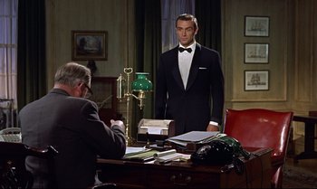 Movie still from “Dr. No” (1962), directed by Terence Young – A man in a tuxedo standing in front of a man in a suit; Medium shot, Over the shoulder angle
