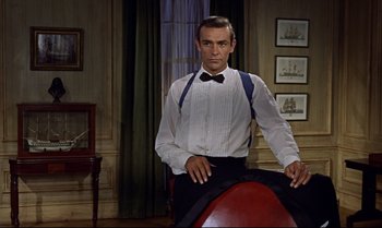 Movie still from “Dr. No” (1962), directed by Terence Young – A man in a white shirt and blue suspenders standing next to a red chair; Medium shot, Low angle