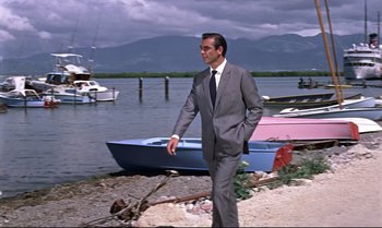 Movie still from “Dr. No” (1962), directed by Terence Young – A man in a suit and tie standing next to a boat; Wide shot, Low angle